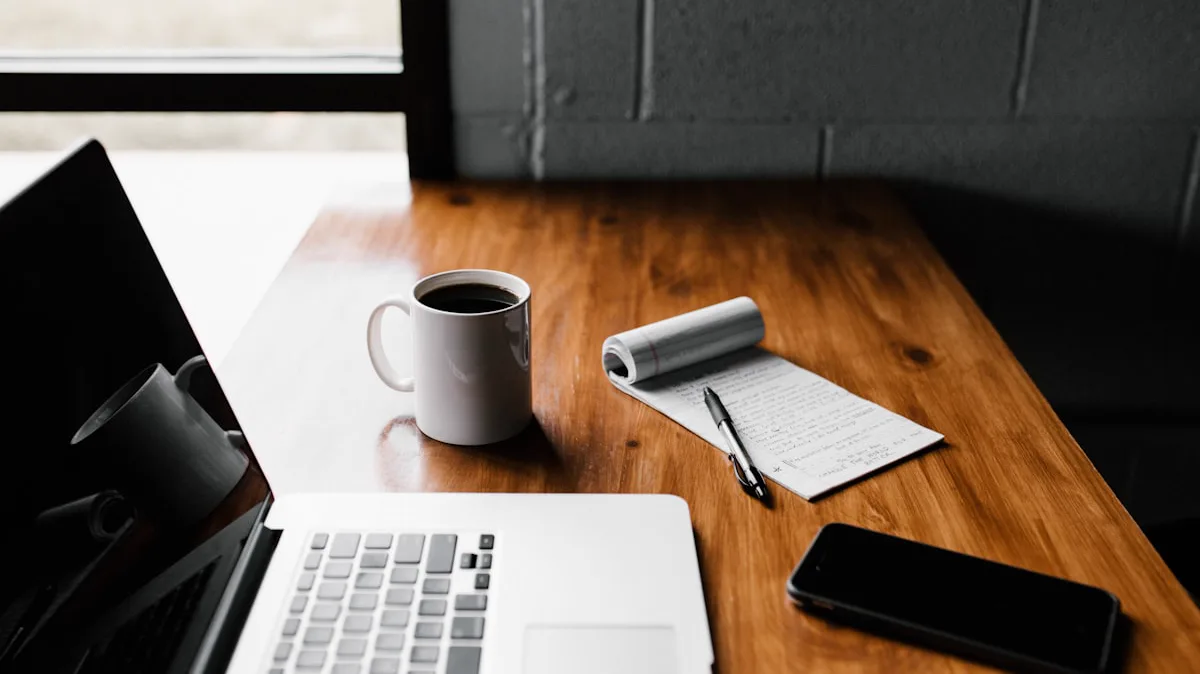 Laptop, cangkir kopi, dan buku catatan berisi tulisan tangan di atas meja kayu
